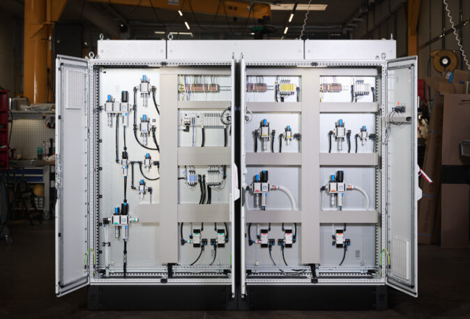 Offene Schalttafeln und Schaltschränke in einer Industriehalle zeigen komplexe Elektro Arbeiten mit Verkabelungssystemen und Komponenten für Mittel- und Hochspannungsanlagen. Die Installation umfasst moderne Glasfaserarbeiten und ist typisch für den Industrie Bereich sowie Anwendungen in Erneuerbaren Energien.