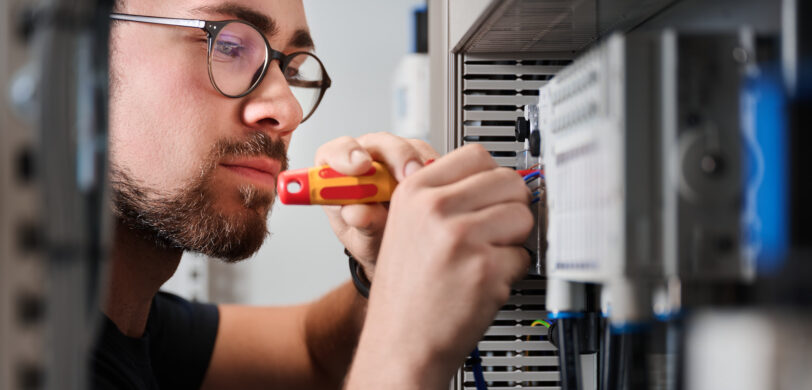 Ein Elektriker führt präzise Elektro Arbeiten an einem Schaltschrank durch, indem er mit einem Schraubendreher Verkabelungssysteme in einer Mittel- und Hochspannungsanlage im Industrie Bereich installiert. Die Arbeit umfasst auch Glasfaserarbeiten und ist Teil moderner Lösungen für Erneuerbare Energien.