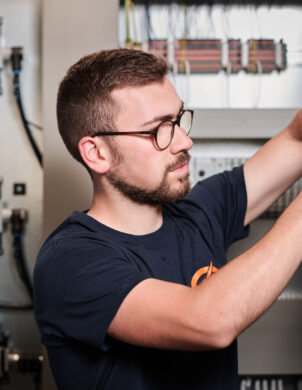 Ein Techniker führt Elektro Arbeiten an einem Schaltschrank durch und verkabelt sorgfältig Mittel- und Hochspannungsanlagen im Industrie Bereich. Im Hintergrund sind Schalttafeln und Verkabelungssysteme zu sehen, die auch Glasfaserarbeiten und Komponenten für Erneuerbare Energien enthalten.