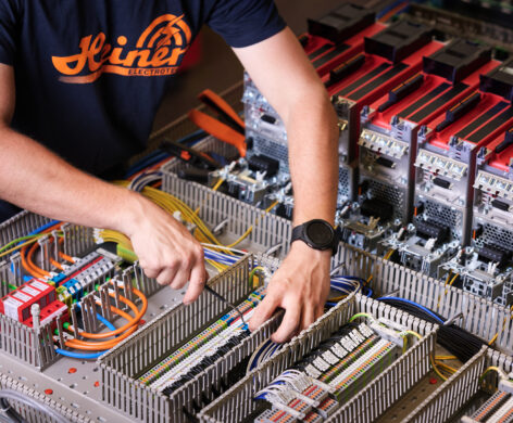 Ein Techniker führt Elektro Arbeiten an einer komplexen Schalttafel mit zahlreichen Kabeln und Schaltschränken durch. Die Verkabelungssysteme sind übersichtlich angeordnet, um Mittel- und Hochspannungsanlagen im Industrie Bereich zu steuern. Im Hintergrund sind Komponenten sichtbar, die auch für Glasfaserarbeiten und Anwendungen im Bereich Erneuerbare Energien genutzt werden können.