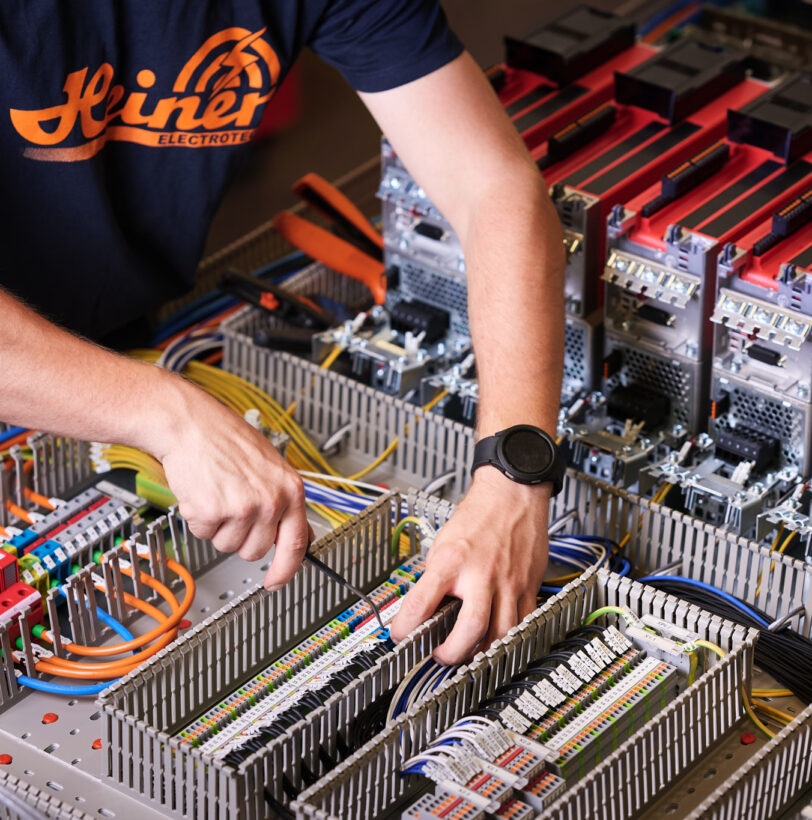 Ein Techniker führt Elektro Arbeiten an einer komplexen Schalttafel mit zahlreichen Kabeln und Schaltschränken durch. Die Verkabelungssysteme sind übersichtlich angeordnet, um Mittel- und Hochspannungsanlagen im Industrie Bereich zu steuern. Im Hintergrund sind Komponenten sichtbar, die auch für Glasfaserarbeiten und Anwendungen im Bereich Erneuerbare Energien genutzt werden können.