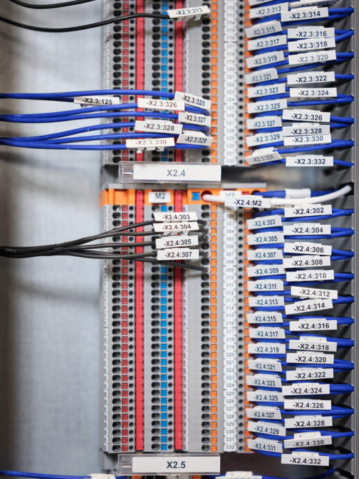 Nahaufnahme einer beschrifteten Verkabelung in einem Schaltschrank mit blauen und schwarzen Kabeln, die in farblich markierte Anschlussleisten eingesteckt sind. Die Kabel sind nummeriert und geordnet, was typische Elektro Arbeiten im Industrie Bereich und bei Mittel- und Hochspannungsanlagen zeigt. Diese Verkabelungssysteme sind essenziell für die Funktion von Schalttafeln und Schaltschränken, auch im Kontext von Glasfaserarbeiten und Erneuerbaren Energien.