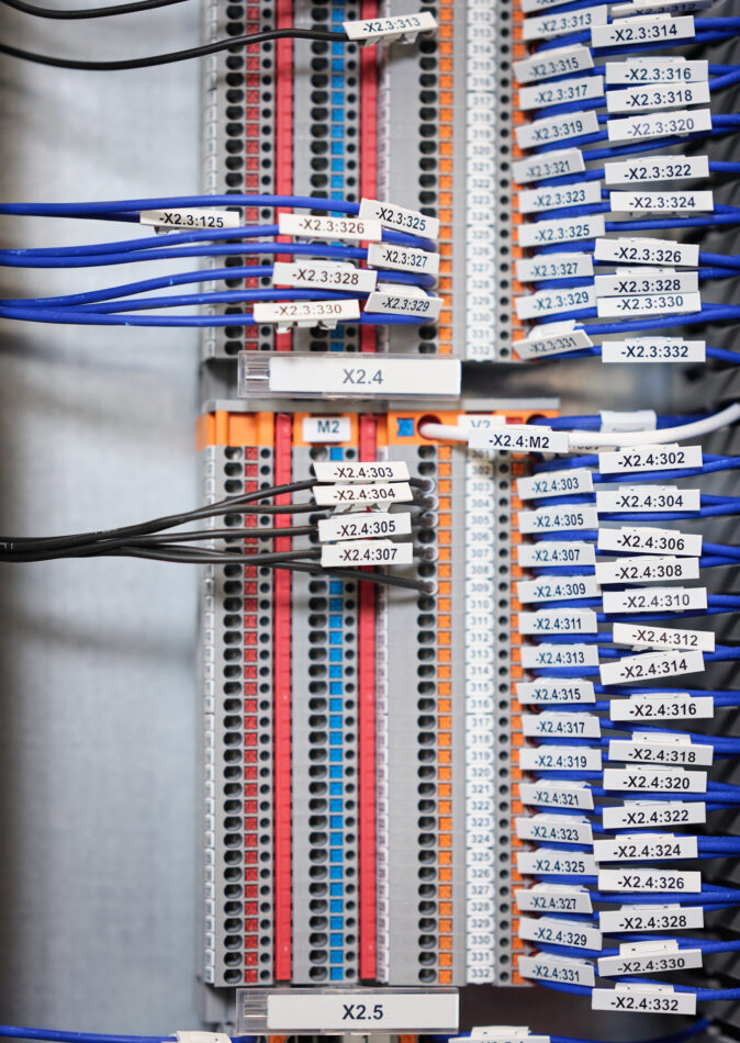 Nahaufnahme einer beschrifteten Verkabelung in einem Schaltschrank mit blauen und schwarzen Kabeln, die in farblich markierte Anschlussleisten eingesteckt sind. Die Kabel sind nummeriert und geordnet, was typische Elektro Arbeiten im Industrie Bereich und bei Mittel- und Hochspannungsanlagen zeigt. Diese Verkabelungssysteme sind essenziell für die Funktion von Schalttafeln und Schaltschränken, auch im Kontext von Glasfaserarbeiten und Erneuerbaren Energien.