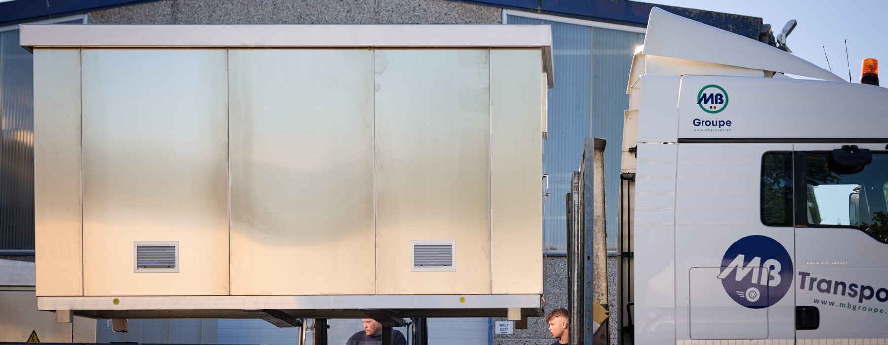 Auf einem Lastwagen wird eine vormontierte Überwachungskabine aus Metall transportiert, die als witterungsbeständige Sicherheitskabine mit thermischer Isolierung dient. Die anschlussfertige Kabine ist ein Beispiel für den Kabinenbau für Bahntechnik und gehört zur Infrabel Loge PNBA. Sie wurde im Rahmen von Prototypenbau und Serienfertigung hergestellt.