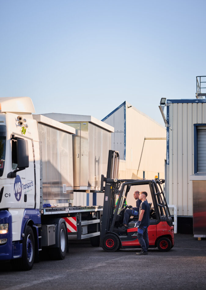 Zwei Arbeiter verladen eine vormontierte Überwachungskabine aus Edelstahl mit thermischer Isolierung auf einen LKW. Die witterungsbeständige Sicherheitskabine ist Teil des Kabinenbaus für Bahntechnik und wird im Prototypenbau und Serienfertigung hergestellt. Die anschlussfertigen Kabinen sind speziell für die Infrabel Loge PNBA konzipiert.