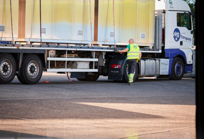 Ein LKW mit einer vormontierten Überwachungskabine wird von einem Mitarbeiter in Warnweste gesichert. Die Infrabel Loge PNBA ist eine witterungsbeständige Sicherheitskabine mit thermischer Isolierung, die speziell im Kabinenbau für Bahntechnik eingesetzt wird. Dieses Modell steht für Prototypenbau und Serienfertigung und ist als anschlussfertige Kabine konzipiert.