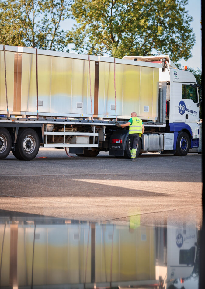 Ein LKW mit einer vormontierten Überwachungskabine wird von einem Mitarbeiter in Warnweste gesichert. Die Infrabel Loge PNBA ist eine witterungsbeständige Sicherheitskabine mit thermischer Isolierung, die speziell im Kabinenbau für Bahntechnik eingesetzt wird. Dieses Modell steht für Prototypenbau und Serienfertigung und ist als anschlussfertige Kabine konzipiert.