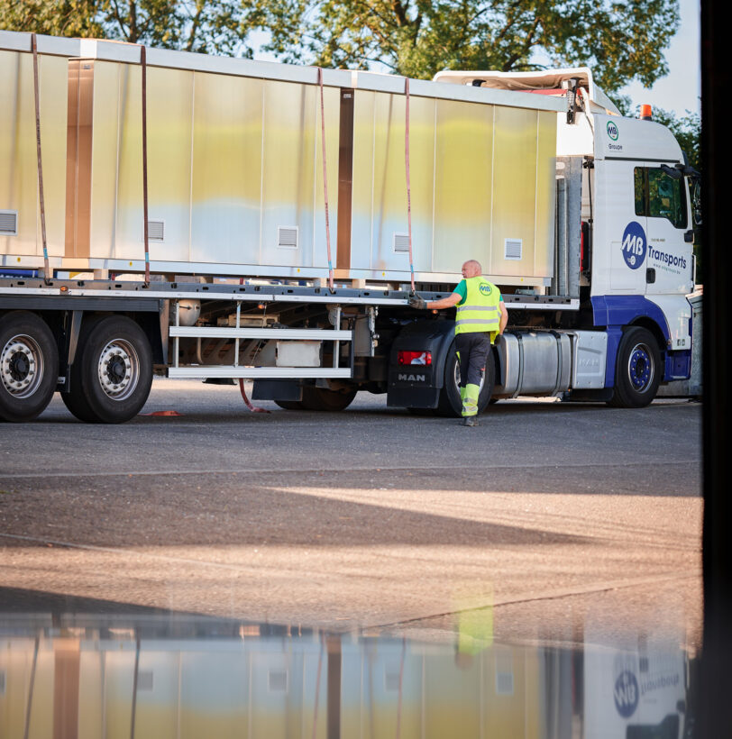 Ein LKW mit einer vormontierten Überwachungskabine wird von einem Mitarbeiter in Warnweste gesichert. Die Infrabel Loge PNBA ist eine witterungsbeständige Sicherheitskabine mit thermischer Isolierung, die speziell im Kabinenbau für Bahntechnik eingesetzt wird. Dieses Modell steht für Prototypenbau und Serienfertigung und ist als anschlussfertige Kabine konzipiert.