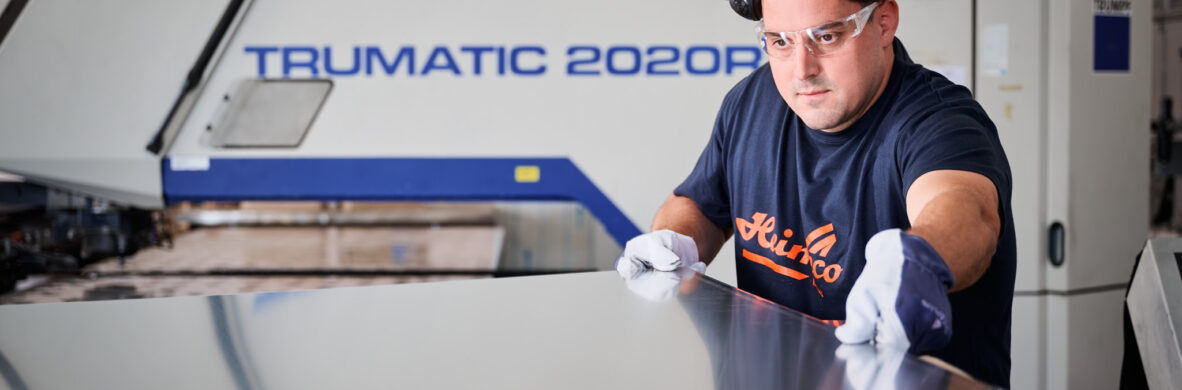 Ein erfahrener Mitarbeiter im Metallbau kontrolliert sorgfältig ein großes Blech in der Fertigungshalle. Er trägt Schutzbrille, Handschuhe und Gehörschutz, während er präzise Maßarbeit bei der Blechverarbeitung leistet. Im Hintergrund ist eine moderne TRUMATIC 2020R Maschine zu sehen, die für Planung, Konstruktion und Produktion von hochwertigen Metallbauteilen steht. Die Kombination aus Qualität und Erfahrung garantiert eine professionelle Fertigung.