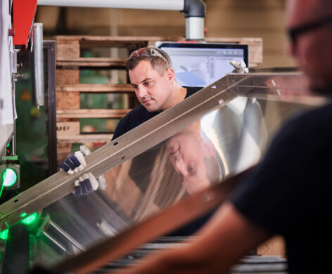 Zwei Facharbeiter im Metallbau prüfen und verarbeiten ein großes Blechstück an einer Maschine. Die Maßarbeit erfordert präzise Planung, Konstruktion und Fertigung. Die erfahrenen Mitarbeiter gewährleisten durch ihre sorgfältige Blechverarbeitung höchste Qualität in der Produktion.