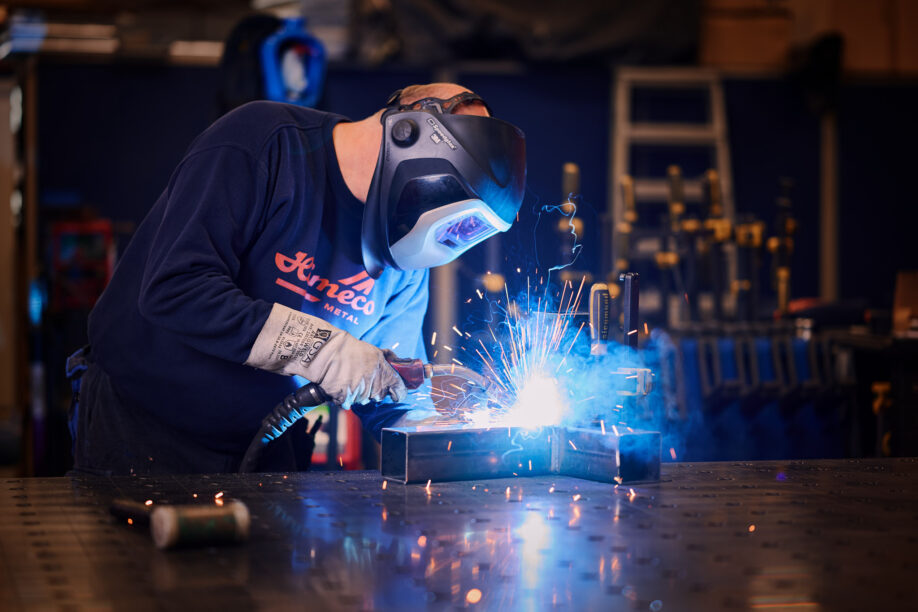 Ein erfahrener Metallbauer führt mit Schutzhelm und Handschuhen präzise Schweißarbeiten an einem Metallrahmen durch. Die Maßarbeit im Metallbau zeigt sich in der sorgfältigen Blechverarbeitung und der exakten Fertigung. Die Planung, Konstruktion und Produktion hochwertiger Metallteile stehen für Qualität und langjährige Erfahrung in der Branche.