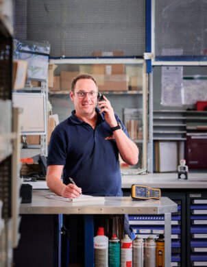 Ein zentraler Ansprechpartner steht hinter einem Arbeitstisch in einer Werkstatt und führt ein Telefongespräch. Er hält einen Stift und notiert etwas auf einem Block, was eine durchdachte und lösungsorientierte Beratung symbolisiert. Im Hintergrund sind Regale mit diversen Werkzeugen und Materialien zu sehen, die die vereinte Kompetenz und Vielseitigkeit des Arbeitsumfelds transparent machen.