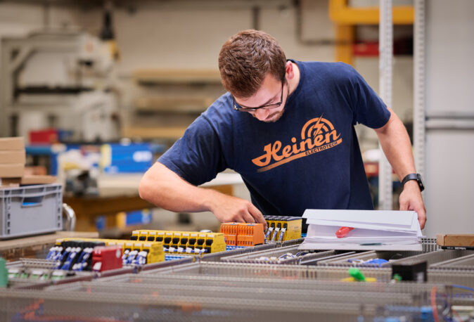 Ein Techniker führt präzise Elektro Arbeiten an Schalttafeln und Schaltschränken in einer Werkstatt durch. Die Verkabelungssysteme sind Teil von Mittel- und Hochspannungsanlagen im Industrie Bereich. Im Hintergrund sind weitere Komponenten und Werkzeuge sichtbar, die auch für Glasfaserarbeiten und Anwendungen im Bereich Erneuerbare Energien genutzt werden.