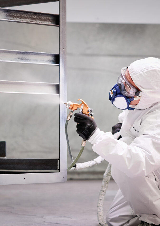 Ein Facharbeiter im Metallbau trägt in Schutzkleidung und Atemmaske eine Lackierung auf eine Metallkonstruktion auf. Die Maßarbeit und präzise Blechverarbeitung sind Teil der sorgfältigen Planung, Konstruktion und Produktion. Die Fertigung erfolgt mit hoher Qualität und langjähriger Erfahrung in der Metallverarbeitung.