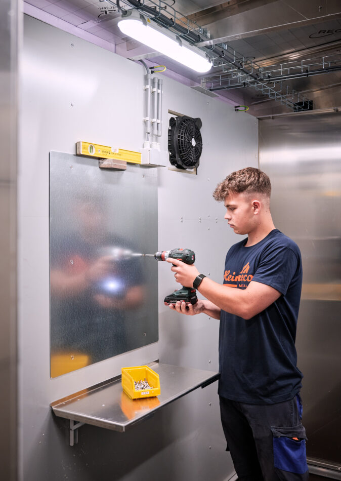 Ein junger Handwerker montiert mit einem Akkuschrauber eine Metallplatte an einer Wand in einer Werkstatt. Die Szene zeigt präzise Maßarbeit im Metallbau mit Fokus auf Blechverarbeitung. Im Vordergrund steht eine gelbe Box mit Schrauben auf einem kleinen, an der Wand befestigten Metalltisch. Die Umgebung vermittelt eine professionelle Planung, Konstruktion und Fertigung von Metallteilen mit hoher Qualität und jahrelanger Erfahrung.