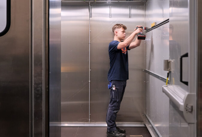 Ein Facharbeiter montiert mit einem Akkuschrauber präzise Metallprofile in einem Raum aus Edelstahlblechen. Die Maßarbeit und Blechverarbeitung erfordert sorgfältige Planung, Konstruktion und Fertigung, um höchste Qualität zu gewährleisten. Die Erfahrung des Metallbauers zeigt sich in der exakten Ausführung und der professionellen Arbeitsweise innerhalb der Metallbauumgebung.