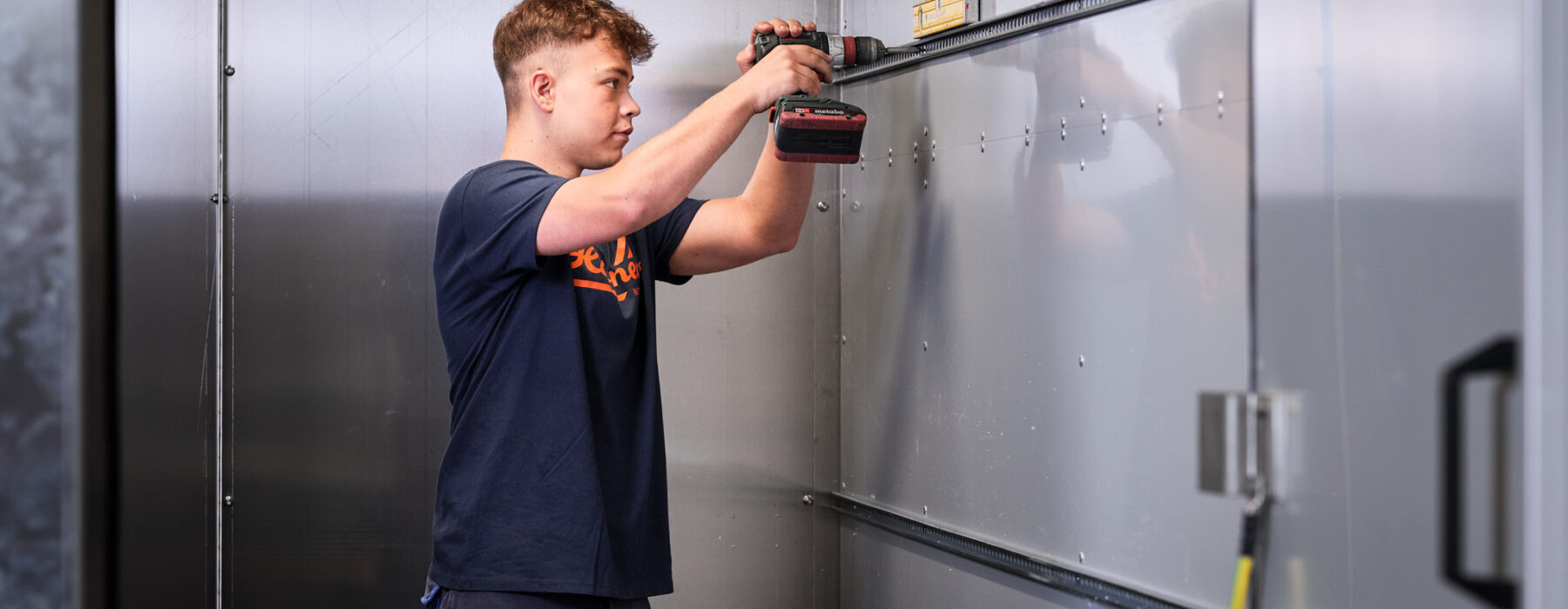 Ein junger Facharbeiter montiert mit einem Akkuschrauber präzise Metallbleche in einem engen Raum. Die Maßarbeit und sorgfältige Blechverarbeitung sind Teil der Planung, Konstruktion und Fertigung im Metallbau. Die hohe Qualität und langjährige Erfahrung spiegeln sich in der professionellen Produktion wider.