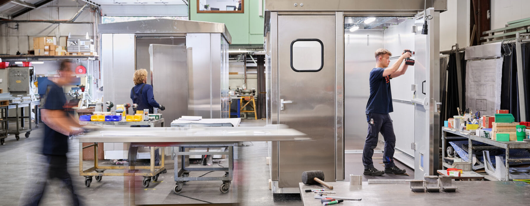 In einer modernen Metallbau-Werkstatt arbeiten Fachkräfte mit hoher Erfahrung an der Maßarbeit von Edelstahlkonstruktionen. Die Blechverarbeitung erfolgt präzise in der Produktion und Fertigung, unterstützt durch sorgfältige Planung und Konstruktion. Ein Mitarbeiter montiert mit einem Akkuschrauber Bauteile an einer Edelstahlkabine, während andere Werkstücke und Werkzeuge übersichtlich in der Werkstatt angeordnet sind. Die Qualität der Arbeit spiegelt sich in der sauberen und organisierten Arbeitsumgebung wider.
