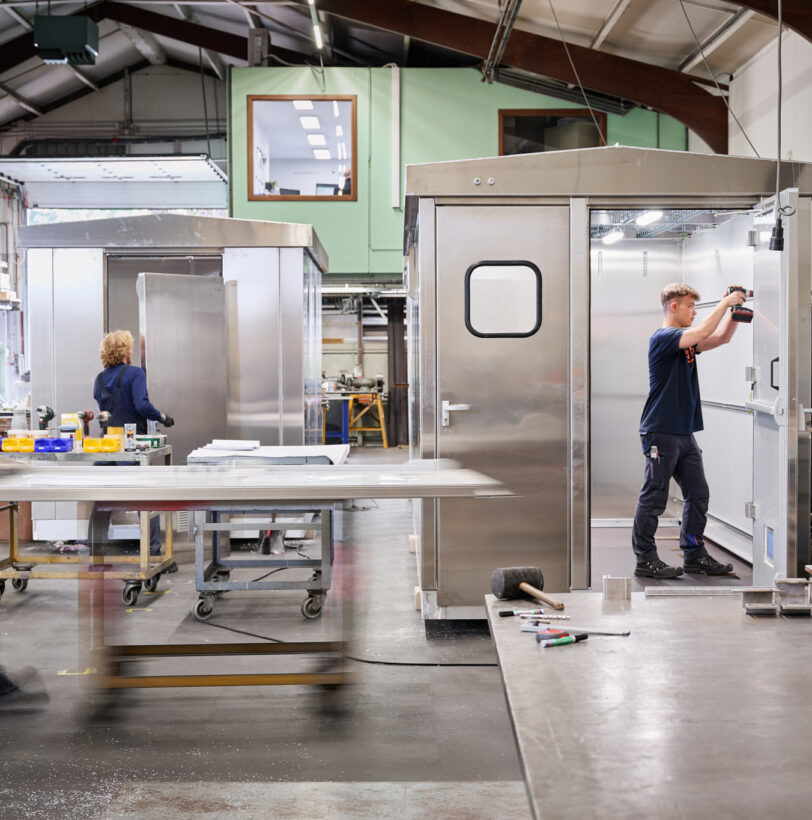 In einer modernen Metallbau-Werkstatt arbeiten Fachkräfte mit hoher Erfahrung an der Maßarbeit von Edelstahlkonstruktionen. Die Blechverarbeitung erfolgt präzise in der Produktion und Fertigung, unterstützt durch sorgfältige Planung und Konstruktion. Ein Mitarbeiter montiert mit einem Akkuschrauber Bauteile an einer Edelstahlkabine, während andere Werkstücke und Werkzeuge übersichtlich in der Werkstatt angeordnet sind. Die Qualität der Arbeit spiegelt sich in der sauberen und organisierten Arbeitsumgebung wider.