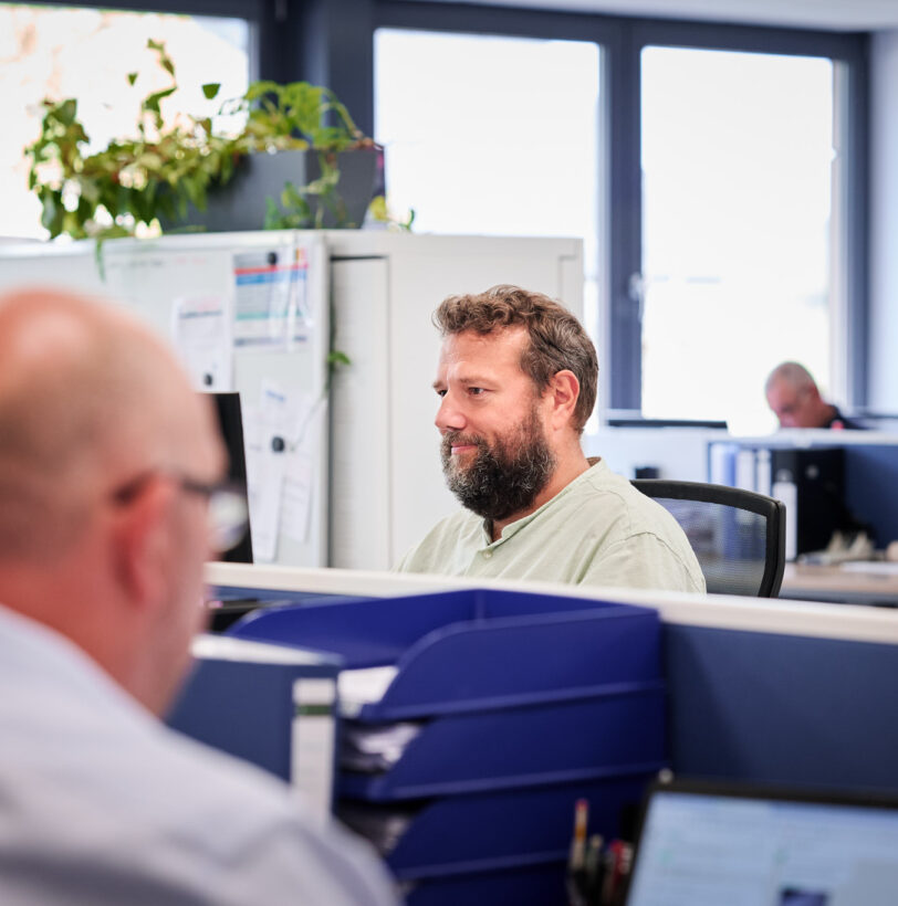 In einem modernen Büro arbeiten mehrere Mitarbeiter konzentriert an ihren Computern. Der zentrale Ansprechpartner mit Kompetenz sitzt durchdacht am Arbeitsplatz und nutzt transparente Dokumentation für die Planung und Entwicklung von Plänen im Projektmanagement. Die Atmosphäre wirkt lösungsorientiert und strukturiert.