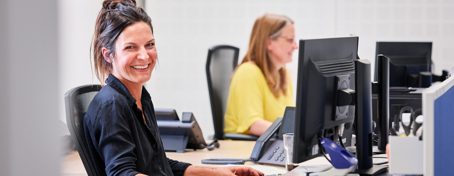 Zwei Frauen arbeiten in einem modernen Büro an Computern. Die vordere Frau lächelt freundlich in die Kamera und symbolisiert den zentralen Ansprechpartner. Das Bild vermittelt vereinte Kompetenz und eine durchdachte, transparente Arbeitsweise. Die vielseitige und lösungsorientierte Beratung spiegelt sich in der professionellen Atmosphäre und der Zusammenarbeit wider.