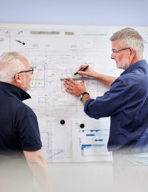 Zwei erfahrene Fachleute besprechen durchdacht und lösungsorientiert Pläne an einer Wandtafel. Der zentrale Ansprechpartner erklärt mit Kompetenz die Details der Projektplanung und Dokumentation im transparenten Projektmanagement.