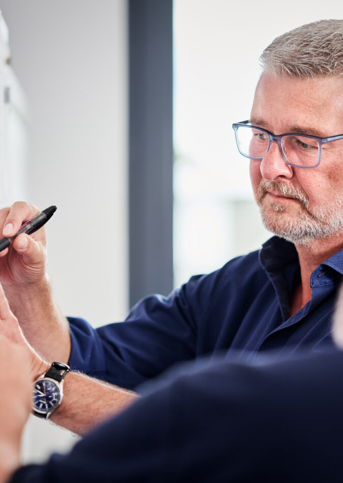 Zwei erfahrene Männer mit Kompetenz arbeiten gemeinsam an durchdachten Plänen an einer Wand. Der zentrale Ansprechpartner erklärt transparent und lösungsorientiert die Dokumentation im Projektmanagement, um eine effiziente Planung und Entwicklung sicherzustellen.