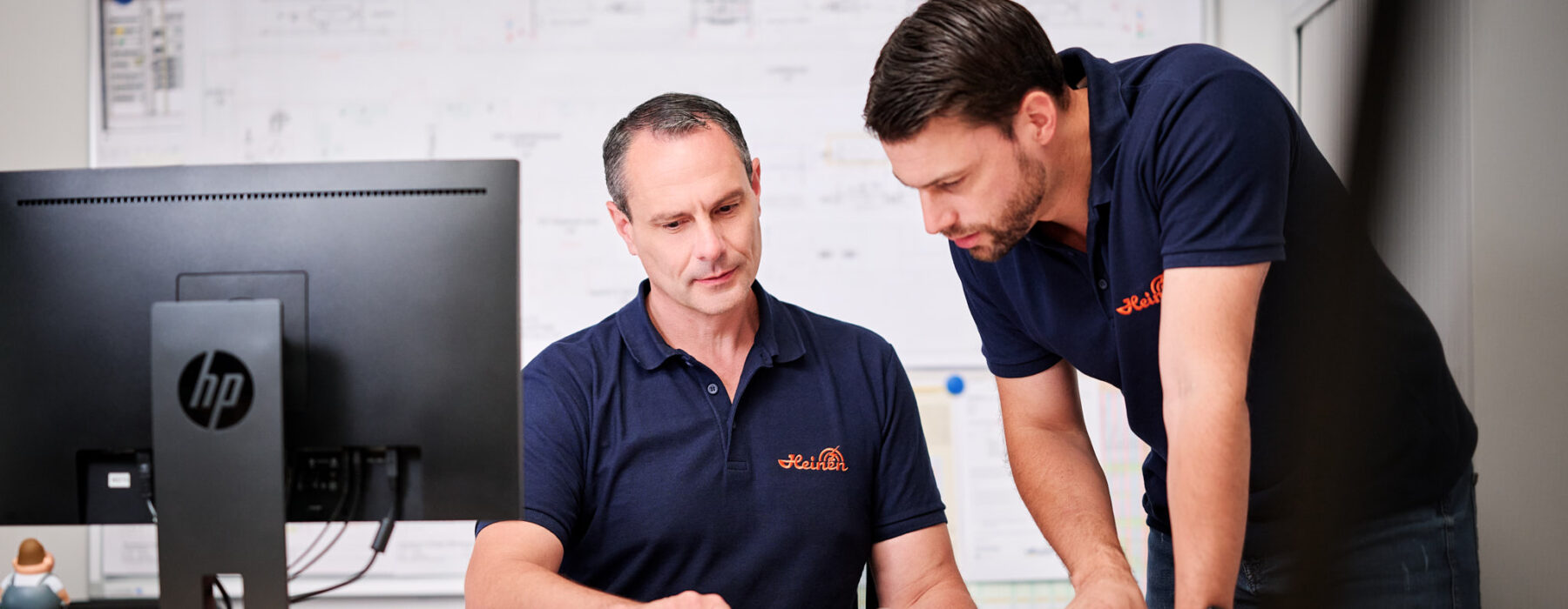 Zwei Männer in dunkelblauen Poloshirts arbeiten gemeinsam an der Planung und Entwicklung eines Projekts. Der zentrale Ansprechpartner erklärt durchdacht die Pläne und Dokumentation, während der andere aufmerksam zuhört. Im Hintergrund sind technische Zeichnungen sichtbar, die ein transparentes und lösungsorientiertes Projektmanagement unterstreichen. Die Kompetenz der beiden zeigt sich in der konzentrierten Zusammenarbeit am Schreibtisch mit Computer und Ordnern.
