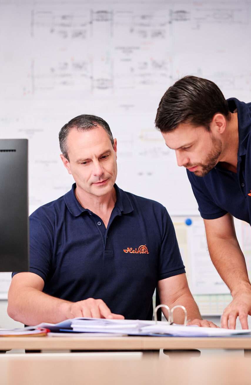 Zwei Männer in dunkelblauen Poloshirts arbeiten gemeinsam an der Planung und Entwicklung eines Projekts. Der zentrale Ansprechpartner erklärt durchdacht die Pläne und Dokumentation, während der andere aufmerksam zuhört. Im Hintergrund sind technische Zeichnungen sichtbar, die ein transparentes und lösungsorientiertes Projektmanagement unterstreichen. Die Kompetenz der beiden zeigt sich in der konzentrierten Zusammenarbeit am Schreibtisch mit Computer und Ordnern.