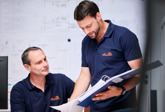 Zwei Männer besprechen Pläne und Dokumentation im Büro mit technischem Hintergrund