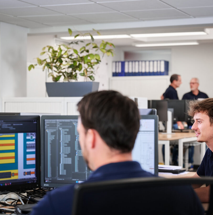 In einem modernen Büro arbeiten mehrere Mitarbeiter konzentriert an Computern mit technischen Plänen und Dokumentationen. Im Hintergrund stehen drei Personen im Gespräch, die als zentraler Ansprechpartner für Projektmanagement und Entwicklung fungieren. Die durchdachte und transparente Planung sowie die lösungsorientierte Kompetenz des Teams sind deutlich erkennbar.