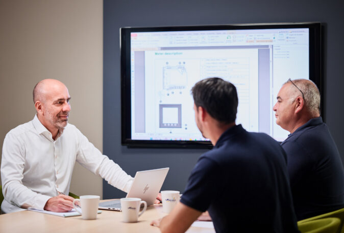 Drei Männer sitzen in einem Besprechungsraum bei einer Projektmanagement-Sitzung. Einer der Teilnehmer, der zentrale Ansprechpartner, präsentiert durchdachte Pläne und Dokumentation auf einem großen Bildschirm. Die Szene zeigt transparente Kommunikation und lösungsorientierte Entwicklung mit hoher Kompetenz und sorgfältiger Planung.