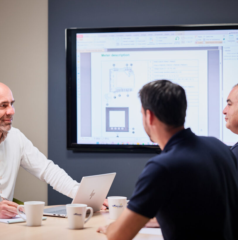 Drei Männer sitzen in einem Besprechungsraum bei einer Projektmanagement-Sitzung. Einer der Teilnehmer, der zentrale Ansprechpartner, präsentiert durchdachte Pläne und Dokumentation auf einem großen Bildschirm. Die Szene zeigt transparente Kommunikation und lösungsorientierte Entwicklung mit hoher Kompetenz und sorgfältiger Planung.