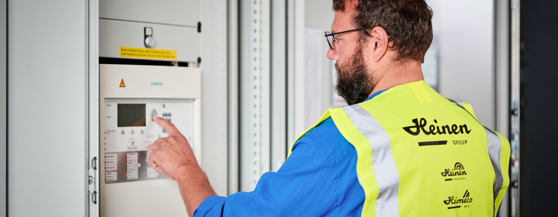Ein Techniker mit Warnweste der Heinen Group bedient eine Siemens-Schaltanlage in einer Industriehalle. Die Aufnahme zeigt die präzise Steuerung von Schaltschränken und Unterverteilungen im Industrieneubau des NMC Neue Hallen Eynatten. Das Bild symbolisiert die Elektroinstallation Großprojekt mit Schwerpunkt auf Schaltanlagenbau, Mittelspannungsstation aus Beton mit Trafo sowie Installation Hauptverteiler mit Unterverteilungen. Weitere Aspekte sind die Niederspannungsverteilung, Brandmeldeanlagen, Industriebeleuchtung LED, Daten- und IT-Verkabelung Produktion und der Anschluss von Produktionslinien.