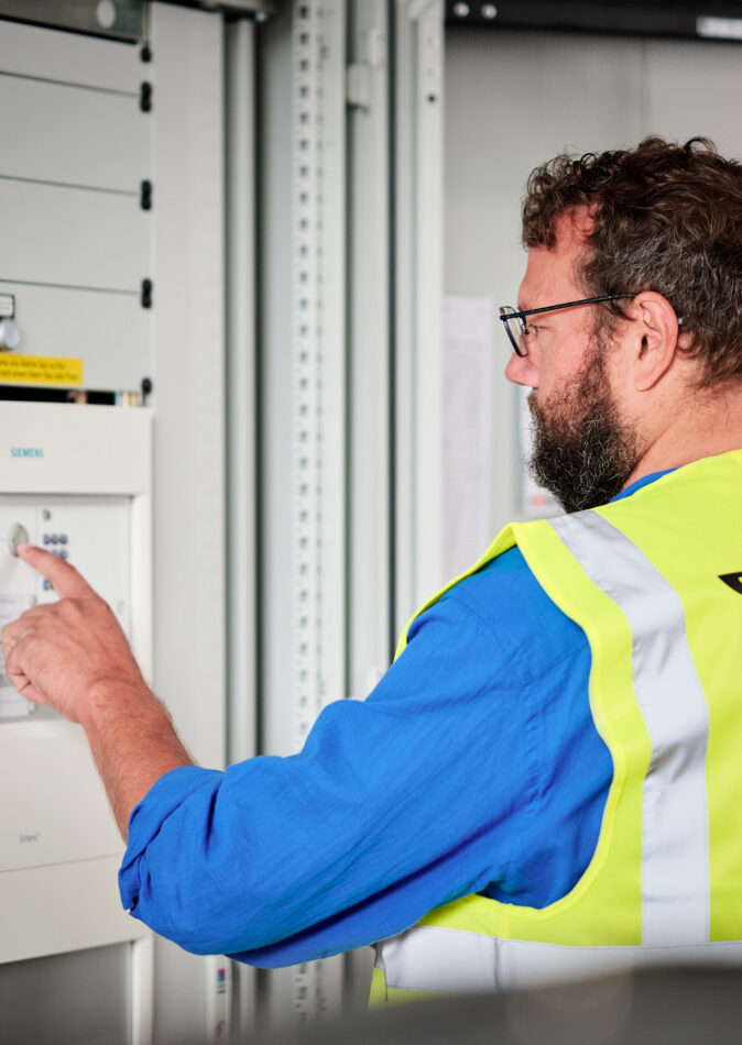 Ein Techniker mit Warnweste der Heinen Group bedient eine Siemens-Schaltanlage in einer Industriehalle. Die Aufnahme zeigt die präzise Steuerung von Schaltschränken und Unterverteilungen im Industrieneubau des NMC Neue Hallen Eynatten. Das Bild symbolisiert die Elektroinstallation Großprojekt mit Schwerpunkt auf Schaltanlagenbau, Mittelspannungsstation aus Beton mit Trafo sowie Installation Hauptverteiler mit Unterverteilungen. Weitere Aspekte sind die Niederspannungsverteilung, Brandmeldeanlagen, Industriebeleuchtung LED, Daten- und IT-Verkabelung Produktion und der Anschluss von Produktionslinien.