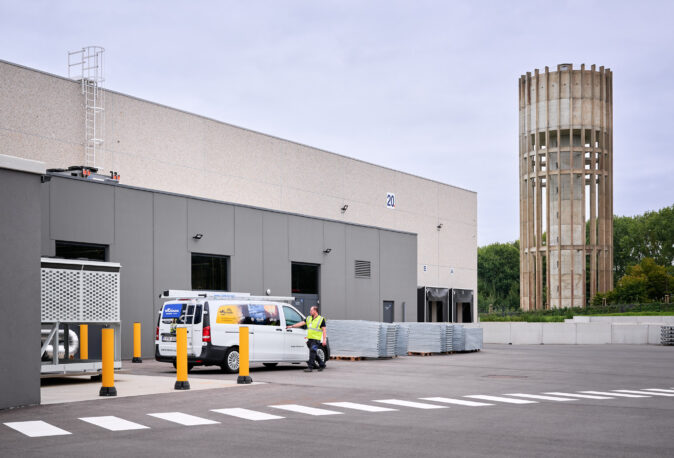 Das Bild zeigt die NMC Neue Hallen Eynatten mit einem grauen Industriegebäude und einem angrenzenden, hohen Beton-Wasserturm. Vor dem Gebäude steht ein weißer Transporter mit der Aufschrift Heinen, daneben ein Arbeiter in Warnweste. Im Außenbereich sind gelbe Schutzpoller und gestapelte Metallplatten zu sehen. Das Bild symbolisiert Elektrotechnik Industriehallen, Schaltschränke und Unterverteilungen im Industrieneubau sowie Elektroinstallation Großprojekt. Die Mittelspannungsstation aus Beton mit Trafo ist durch den Wasserturm angedeutet. Die Installation Hauptverteiler mit Unterverteilungen, Niederspannungsverteilung, Brandmeldeanlagen, Industriebeleuchtung LED sowie Daten- und IT-Verkabelung Produktion und Anschluss Produktionslinien sind typische Bestandteile dieses Industriekomplexes.