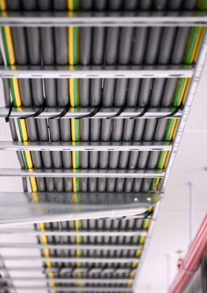 Detailaufnahme der Elektroinstallation in den NMC Neue Hallen Eynatten mit Kabeltrassen für Schaltschränke und Unterverteilungen im Industrieneubau. Zu sehen sind sorgfältig verlegte Leitungen für Niederspannungsverteilung, Daten- und IT-Verkabelung Produktion sowie Installationen für Brandmeldeanlagen und Industriebeleuchtung LED. Die Installation ist Teil eines Elektroinstallation Großprojekts inklusive Schaltanlagenbau und Anschluss Produktionslinien.