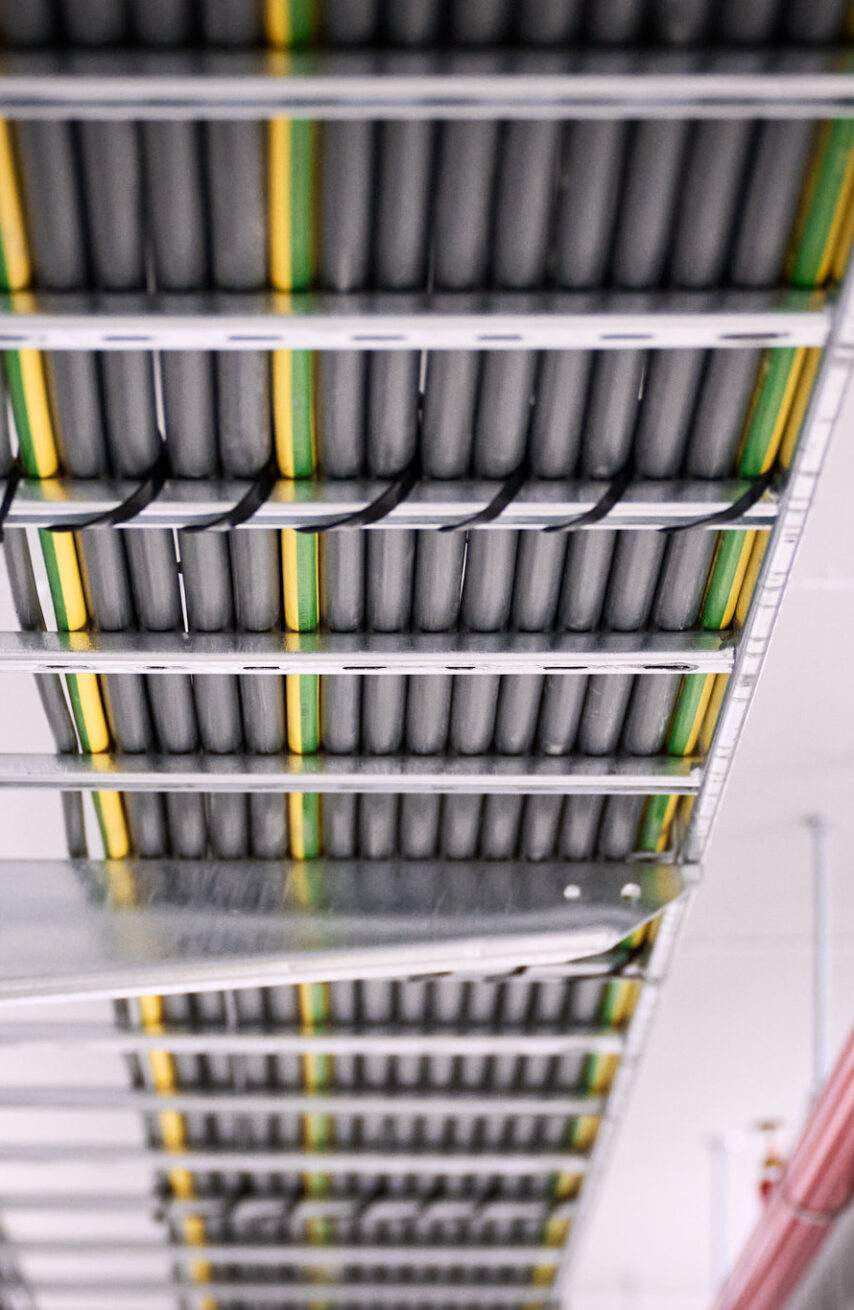 Detailaufnahme der Elektroinstallation in den NMC Neue Hallen Eynatten mit Kabeltrassen für Schaltschränke und Unterverteilungen im Industrieneubau. Zu sehen sind sorgfältig verlegte Leitungen für Niederspannungsverteilung, Daten- und IT-Verkabelung Produktion sowie Installationen für Brandmeldeanlagen und Industriebeleuchtung LED. Die Installation ist Teil eines Elektroinstallation Großprojekts inklusive Schaltanlagenbau und Anschluss Produktionslinien.