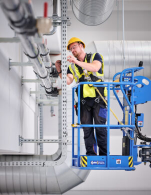 Ein Elektriker arbeitet auf einer Hebebühne in den NMC Neue Hallen Eynatten und installiert Elektrotechnik in Industriehallen. Er montiert Schaltschränke und Unterverteilungen im Industrieneubau, Teil eines Elektroinstallation Großprojekts. Im Hintergrund sind Lüftungsrohre und Kabeltrassen sichtbar, die zur Installation von Schaltanlagenbau und Niederspannungsverteilung gehören. Die Szene zeigt die Installation von Hauptverteilern mit Unterverteilungen, Industriebeleuchtung LED sowie Daten- und IT-Verkabelung für den Anschluss von Produktionslinien. Die Mittelspannungsstation aus Beton mit Trafo ist nicht sichtbar, aber Teil des Gesamtprojekts inklusive Brandmeldeanlagen.