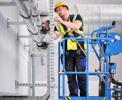 Ein Elektriker arbeitet auf einer Hebebühne in den NMC Neue Hallen Eynatten und installiert Elektrotechnik in Industriehallen. Er montiert Schaltschränke und Unterverteilungen im Industrieneubau, Teil eines Elektroinstallation Großprojekts. Im Hintergrund sind Lüftungsrohre und Kabeltrassen sichtbar, die zur Installation von Schaltanlagenbau und Niederspannungsverteilung gehören. Die Szene zeigt die Installation von Hauptverteilern mit Unterverteilungen, Industriebeleuchtung LED sowie Daten- und IT-Verkabelung für den Anschluss von Produktionslinien. Die Mittelspannungsstation aus Beton mit Trafo ist nicht sichtbar, aber Teil des Gesamtprojekts inklusive Brandmeldeanlagen.
