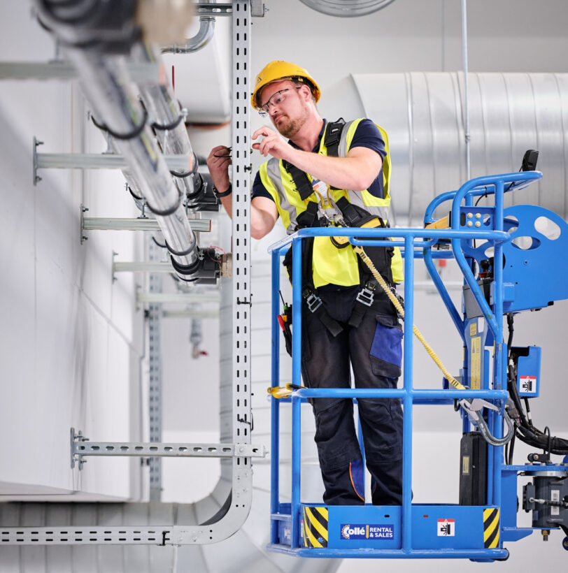 Ein Elektriker arbeitet auf einer Hebebühne in den NMC Neue Hallen Eynatten und installiert Elektrotechnik in Industriehallen. Er montiert Schaltschränke und Unterverteilungen im Industrieneubau, Teil eines Elektroinstallation Großprojekts. Im Hintergrund sind Lüftungsrohre und Kabeltrassen sichtbar, die zur Installation von Schaltanlagenbau und Niederspannungsverteilung gehören. Die Szene zeigt die Installation von Hauptverteilern mit Unterverteilungen, Industriebeleuchtung LED sowie Daten- und IT-Verkabelung für den Anschluss von Produktionslinien. Die Mittelspannungsstation aus Beton mit Trafo ist nicht sichtbar, aber Teil des Gesamtprojekts inklusive Brandmeldeanlagen.