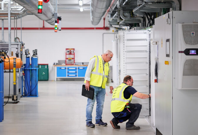 Zwei Techniker prüfen Schaltschränke und Unterverteilungen im Industrieneubau der NMC Neue Hallen Eynatten. Die Elektroinstallation im Großprojekt umfasst Schaltanlagenbau, Mittelspannungsstation aus Beton mit Trafo sowie die Installation von Hauptverteilern mit Unterverteilungen. Im Hintergrund sind Industriebeleuchtung LED, Brandmeldeanlagen und Daten- und IT-Verkabelung für die Produktion sichtbar. Die Techniker sichern den Anschluss der Produktionslinien in der Niederspannungsverteilung ab.