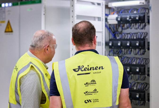 Zwei Techniker in gelben Warnwesten der Heinen Group inspizieren Schaltschränke und Unterverteilungen im Rahmen der Elektroinstallation eines Großprojekts in den NMC Neue Hallen Eynatten. Im Hintergrund sind komplexe Schaltanlagen mit zahlreichen Kabeln und Sicherungen zu sehen, die Teil des Schaltanlagenbaus und der Niederspannungsverteilung sind. Das Bild symbolisiert die professionelle Installation von Hauptverteilern mit Unterverteilungen, Mittelspannungsstationen aus Beton mit Trafo, Brandmeldeanlagen, Industriebeleuchtung LED sowie Daten- und IT-Verkabelung für den Anschluss von Produktionslinien in Industriehallen.