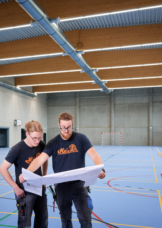 In der Sporthalle Schönefeld Eupen stehen zwei Handwerker mit Bauplänen auf dem blauen Mehrzweck-Sportboden. Die Halle ist barrierefrei gestaltet und verfügt über eine moderne elektrotechnische Ausstattung mit intelligenter LED-Beleuchtung, IT-Verkabelung und Sicherheitstechnik. Die Decke zeigt sichtbare Lüftungsrohre und Holzträger, während große Fenster für Tageslicht sorgen. Im Hintergrund sind Sportgeräte und ein Handballtor zu sehen. Die Halle ist zudem mit einer Photovoltaikanlage zur nachhaltigen Energiegewinnung ausgestattet.