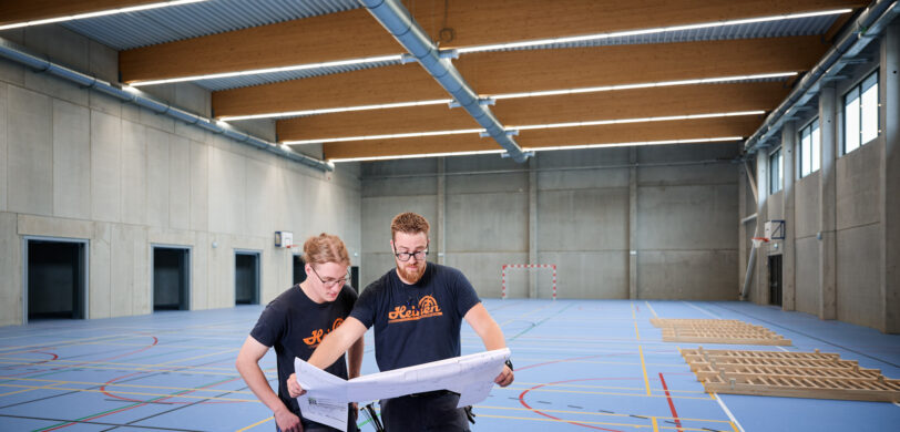 In der Sporthalle Schönefeld Eupen stehen zwei Handwerker mit Bauplänen auf dem blauen Mehrzweck-Sportboden. Die Halle ist barrierefrei gestaltet und verfügt über eine moderne elektrotechnische Ausstattung mit intelligenter LED-Beleuchtung, IT-Verkabelung und Sicherheitstechnik. Die Decke zeigt sichtbare Lüftungsrohre und Holzträger, während große Fenster für Tageslicht sorgen. Im Hintergrund sind Sportgeräte und ein Handballtor zu sehen. Die Halle ist zudem mit einer Photovoltaikanlage zur nachhaltigen Energiegewinnung ausgestattet.