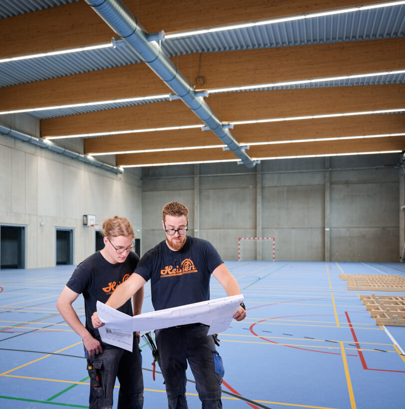 Zwei Handwerker prüfen Baupläne in barrierefreier Sporthalle Schönefeld Eupen mit moderner Elektrotechnik