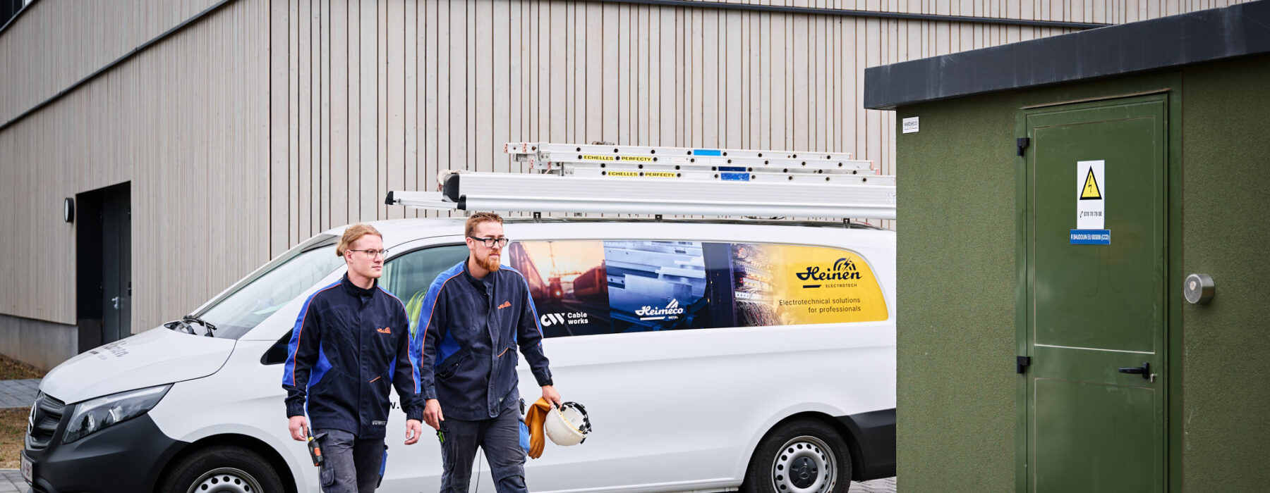 Zwei Elektriker in Arbeitskleidung gehen vor der Sporthalle Schönefeld Eupen, neben einem weißen Firmenfahrzeug mit Leitern auf dem Dach. Die Elektrotechnische Ausstattung der barrierefreien Mehrzweckhalle umfasst unter anderem eine Photovoltaikanlage, intelligente LED-Beleuchtung, IT-Verkabelung und moderne Sicherheitstechnik.