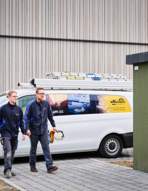 Zwei Elektriker in Arbeitskleidung gehen vor der Sporthalle Schönefeld Eupen, neben einem weißen Firmenfahrzeug mit Leitern auf dem Dach. Die Elektrotechnische Ausstattung der barrierefreien Mehrzweckhalle umfasst unter anderem eine Photovoltaikanlage, intelligente LED-Beleuchtung, IT-Verkabelung und moderne Sicherheitstechnik.