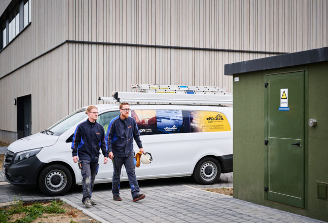 Zwei Elektriker in Arbeitskleidung gehen vor der Sporthalle Schönefeld Eupen, neben einem weißen Firmenfahrzeug mit Leitern auf dem Dach. Die Elektrotechnische Ausstattung der barrierefreien Mehrzweckhalle umfasst unter anderem eine Photovoltaikanlage, intelligente LED-Beleuchtung, IT-Verkabelung und moderne Sicherheitstechnik.