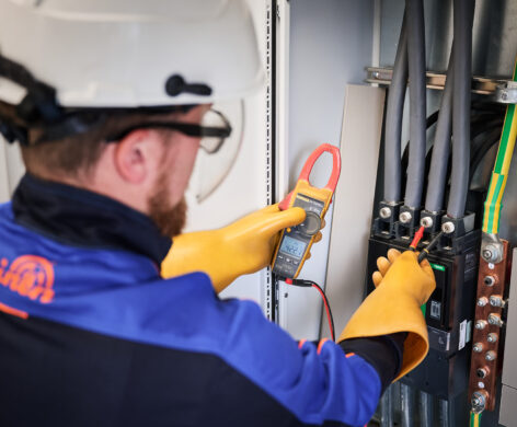 Ein Elektriker in Schutzhelm und Isolierhandschuhen prüft mit einem Stromzangenmessgerät die elektrotechnische Ausstattung eines Schaltschranks. Die Installation gehört zur Sicherheitstechnik und IT-Verkabelung der barrierefreien Mehrzweckhalle in der Sporthalle Schönefeld Eupen, die zudem mit einer Photovoltaikanlage und intelligenter LED-Beleuchtung ausgestattet ist.